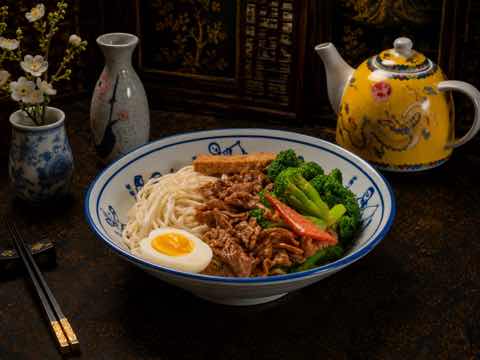 Beef noodle soup with broccoli, egg and greens in a blue and white bowl