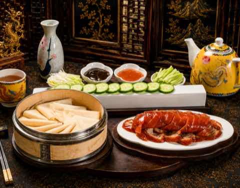Peking duck sliced onto a white platter beside a steamer of warm pancakes, cucumber, spring onion and dipping sauces