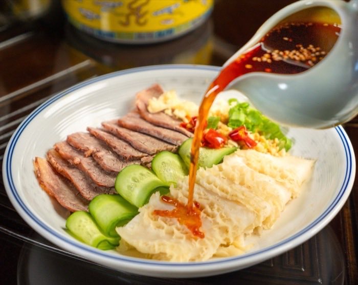 Aromatic sauce being poured over a cold platter of sliced meat and cucumber
