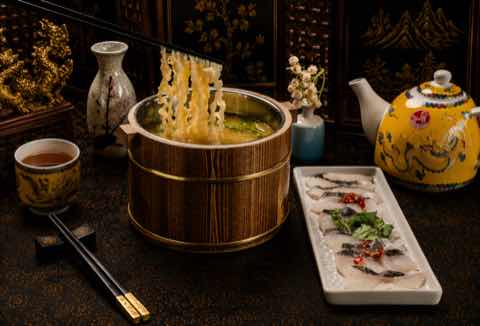 Noodles lifted from a wooden bucket beside a plate of sliced fish