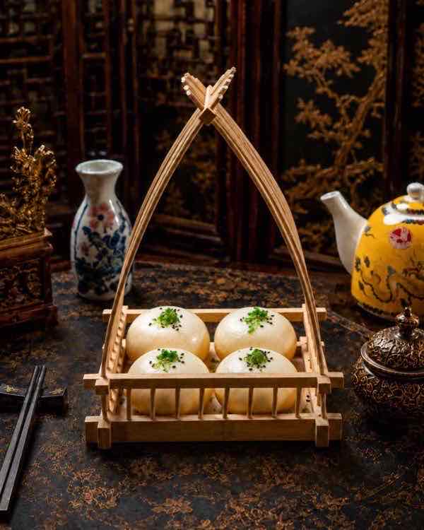 Steamed buns finished with spring onion and black sesame, presented in a handled bamboo carrier beside a dragon-painted teapot on a gold-inlaid lacquer table